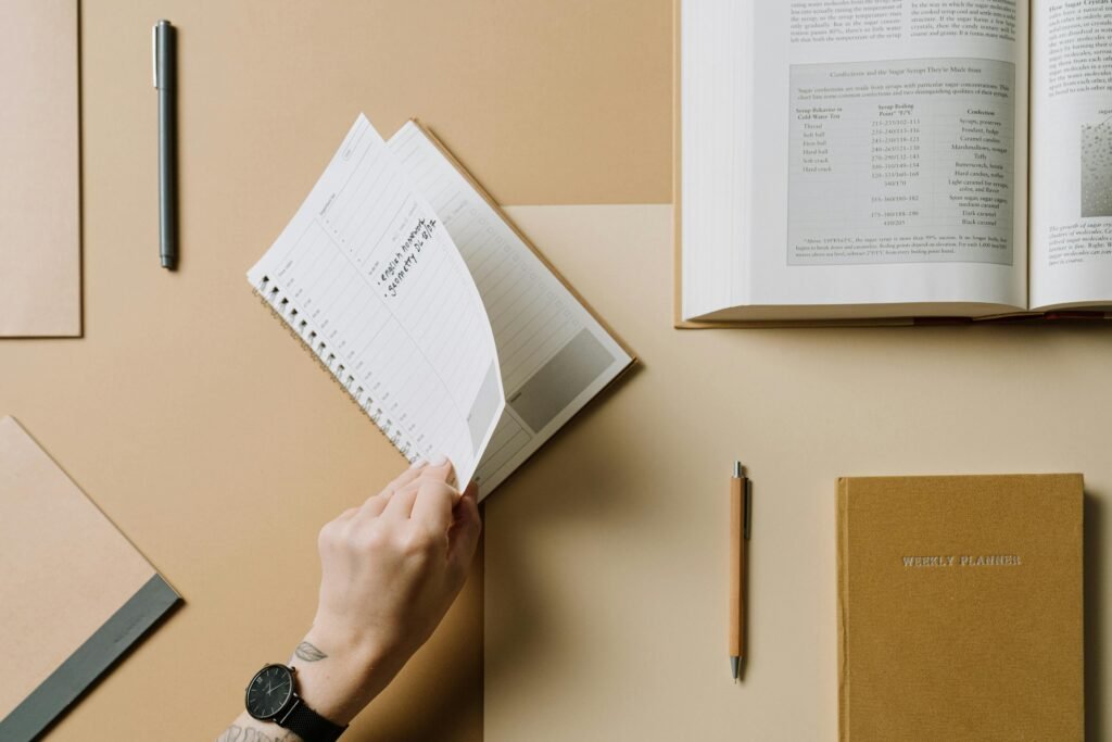 Aerial view of a hand turning pages of a spiral notebook with planner and pens on a desk.