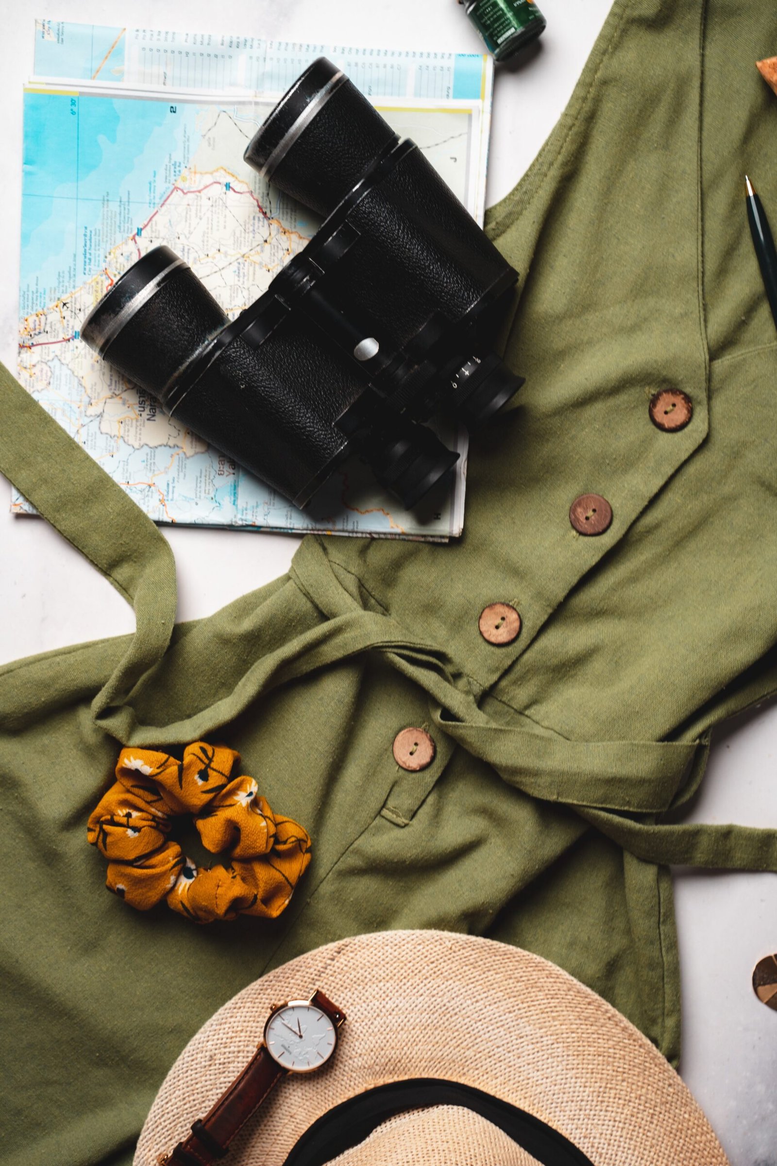 Flat lay of army green dress, binoculars, map, and travel items for adventure planning.